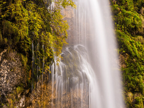 Waterfall cascades down wet mossy rocky cliffs framed by vibrant golden autumn foliage, iconic outdoor nature tourism destination, Goriuda waterfall, Julian Alps, Chiusaforte, Friuli, Italy.
