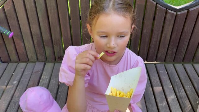 Young girl wearing a pink hat sitting on a park bench enjoying french fries outdoors