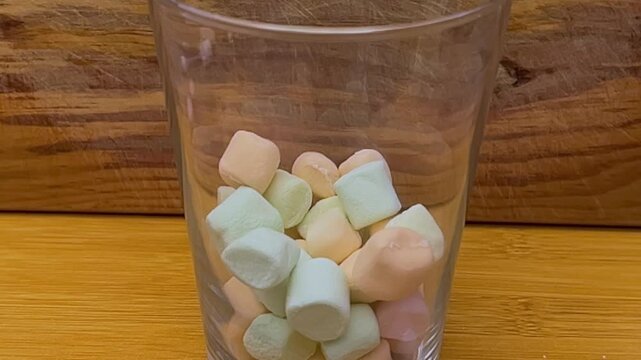 Close up of colorful marshmallows falling into a clear glass on a wooden table, capturing soft candy pieces in motion with a simple indoor composition