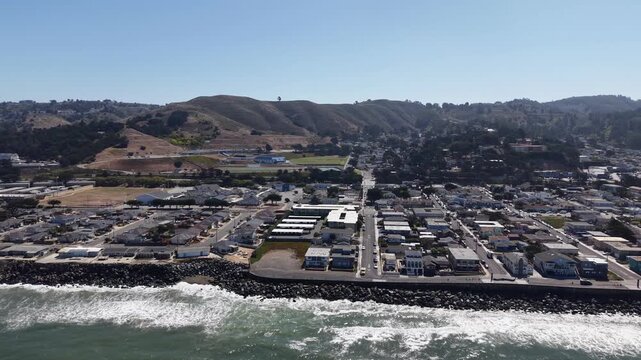 Aerial view of the coastal town of Pacifica, California, showing houses along the Pacific Ocean coastline with rolling hills in the background on a bright, sunny day with clear blue skies