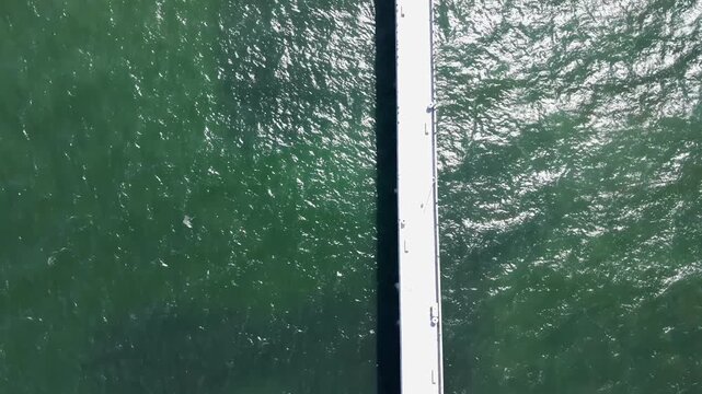 Top down drone footage flying along the white concrete structure of the Pacifica Municipal Pier extending into the green waters of the Pacific Ocean on a sunny morning