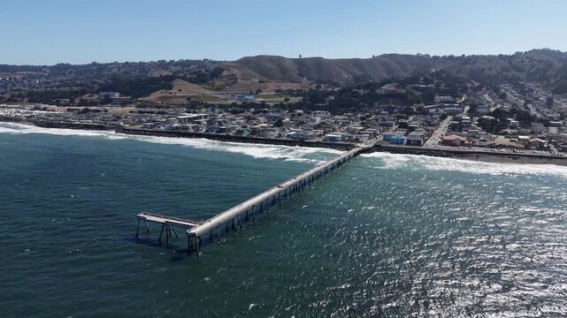 Expansive aerial view showing the Pacifica Municipal Pier extending into the Pacific Ocean, with the coastal city and rolling hills in the background on a bright, sunny day