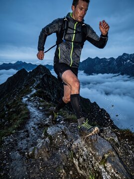 Athletic man trail running along a rocky mountain ridge above the clouds at dusk, splashing through water, symbolizing endurance, fitness and outdoor adventure in dramatic terrain.
