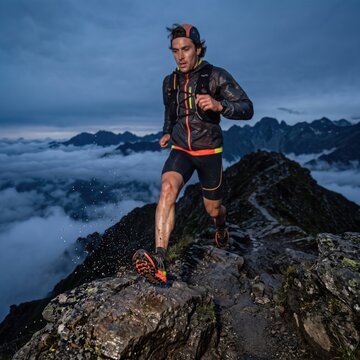 Athletic man trail running along a rocky mountain ridge above the clouds at dusk, splashing through water, symbolizing endurance, fitness and outdoor adventure in dramatic terrain.