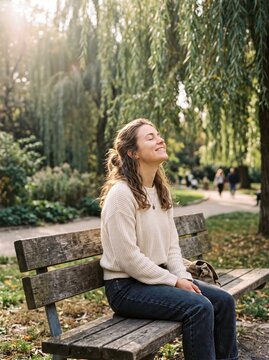 Smiling woman sits on a wooden bench under weeping willow trees, eyes closed and face turned to the sun, enjoying a peaceful moment outdoors, symbolizing relaxation and mindfulness.