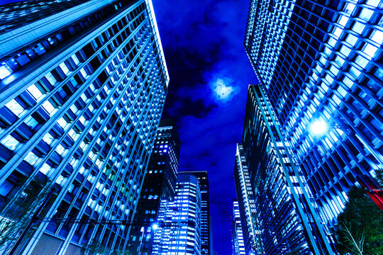 Dynamic low-angle view of illuminated skyscrapers in Tokyo at night under a cloudy sky with glowing full moon.