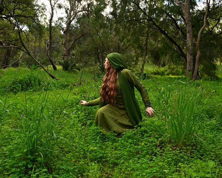 full length portrait of beautiful female model, sitting  kneeling figure picking herb plants, wearing hooded green medieval historical gown costume with cloak. Magical forest background with trees.