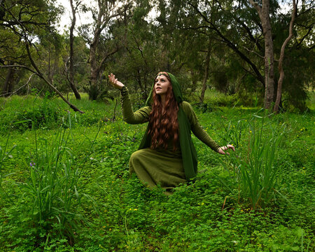 full length portrait of beautiful female model, sitting  kneeling figure picking herb plants, wearing hooded green medieval historical gown costume with cloak. Magical forest background with trees.