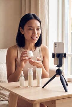 Young woman demonstrating skincare products on a smartphone tripod for social media beauty tutorial and product review background for banner