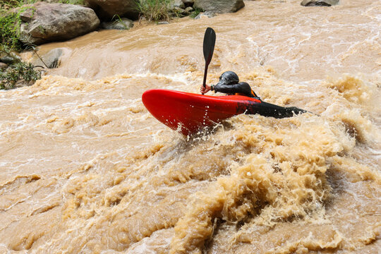 Kayaker navigating muddy whitewater rapids in red kayak, wearing helmet and paddle, dynamic action and adventurous mood