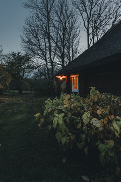Laane County, Estonia - 29 October 2025: View of a traditional wooden house with a glowing window and thatched roof surrounded by autumn foliage and bare trees on Vormsi island.