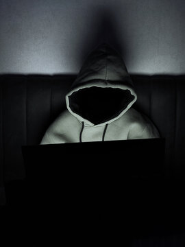 Anonymous person wearing a hooded sweatshirt using a laptop in a dark room at night. Concept of cybersecurity, hacker, online privacy, internet security and anonymous user.
