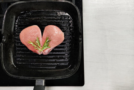 Two raw meat steaks arranged in heart shape on grill pan with rosemary. Top view of fresh uncooked beef with herbs, romantic food concept.
