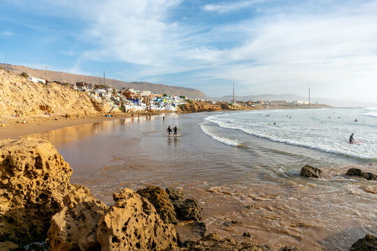 Imsouane, Souss-Massa, Morocco - 11 January 2024: View of surfers walking along the sandy beach with rocky cliffs and coastal buildings in the background under a bright sky.