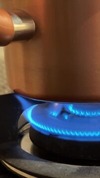Vertical 4K shot of blue gas stove flame heating a nonstick teflon pot to boil water in a kitchen. 