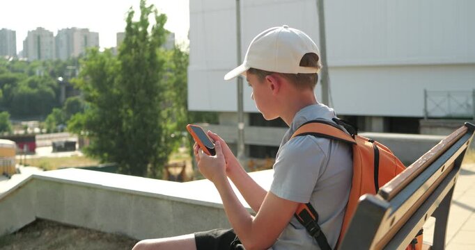 Young boy checking smartphone while sitting on city bench with backpack. Urban scene focuses on digital navigation, location awareness and using mobile technology while exploring city