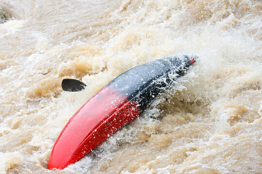 Red and black kayak capsized in turbulent muddy river rapids, paddler partially submerged with paddle blade visible, dynamic action and splashing water conveying danger and excitement
