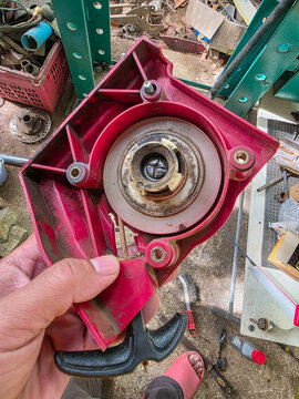 Red plastic recoil starter housing with pull handle and metal pulley assembly showing wear and grease in workshop scene