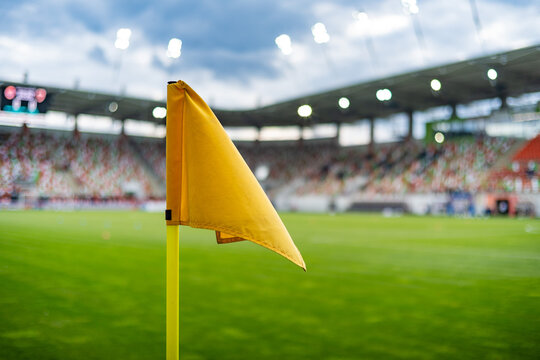 Yellow soccer corner flag on a green football field with blurred stadium and spectators under bright floodlights, creating a colorful sports background.