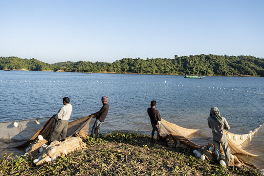 Rangamati, Bangladesh - 14 December 2024: View of fishermen casting nets into the calm waters of Kaptai Lake with lush green hills in the background under a clear blue sky.