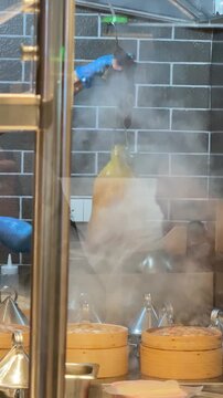 Vertical 4K video of a chef basting hanging whole duck with hot broth next to dim sum bamboo steamers. 