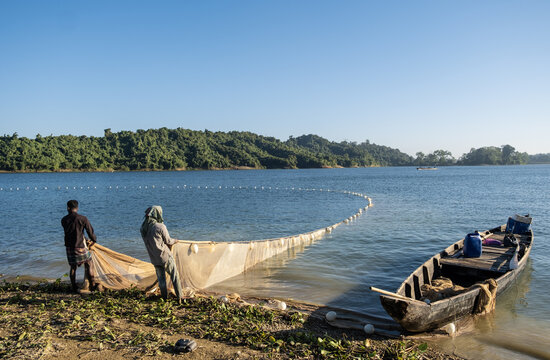 Rangamati, Chittagong Division, Bangladesh - 14 December 2024: View of fishermen pulling a large net from the shore of Kaptai Lake with a wooden boat and lush green hills in the background.