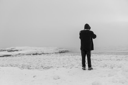 Aerial view of a person in a dark coat and hood standing on a snow-covered beach by a frozen lake under a grey sky, Harrisville, Michigan, United States.