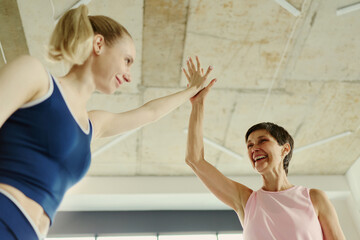 Fototapeta premium Young Caucasian woman and senior Caucasian woman giving high five in gym, celebrating fitness success. Low angle shot useful for active aging and intergenerational wellness campaigns