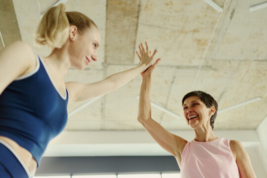 Young Caucasian woman and senior Caucasian woman giving high five in gym, celebrating fitness success. Low angle shot useful for active aging and intergenerational wellness campaigns