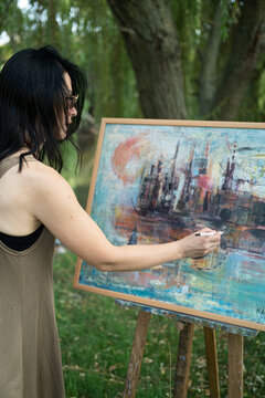 Beautiful woman painter by the river with sunglasses painting on a canvas on an easel, behind view over the shoulder