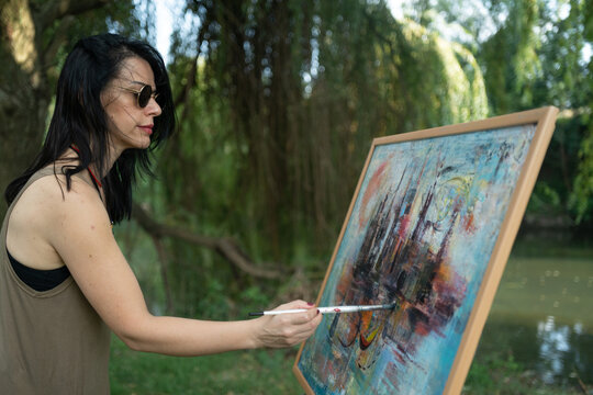 Beautiful woman painter by the river with sunglasses painting on a canvas on an easel