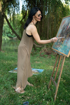 Beautiful woman painter by the river with sunglasses painting on a canvas on an easel