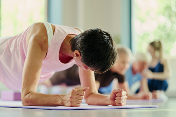 Fototapeta premium Mature Caucasian woman performing forearm plank exercise during group fitness class in gym. Image represents active lifestyle and is suitable for wellness marketing, low angle shot
