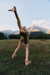 Obraz premium Young girl dances in a sunlit meadow with mountains in the background, arms raised in joy
