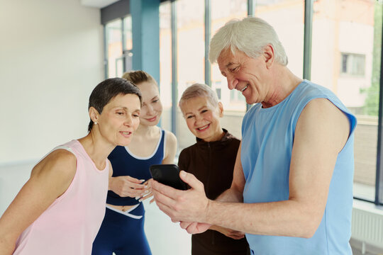 Diverse Caucasian group of senior and young fitness clients looking at smartphone screen in gym. Image represents digital health tracking and active lifestyle marketing