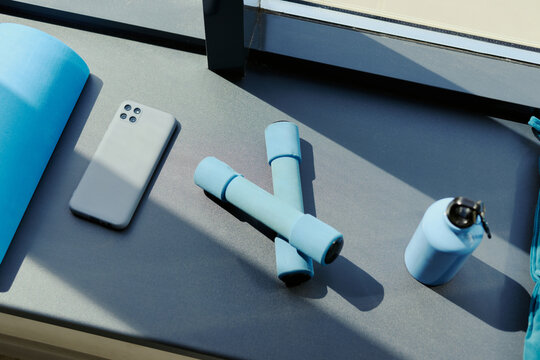 Fitness equipment including dumbbells, water bottle, smartphone, and yoga mat lying on windowsill. High angle view of workout gear representing home training and wellness app marketing