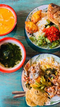 Top down flat lay view of a traditional Indonesian food spread featuring Nasi Tempong with fried fish, sambal chili paste, fresh vegetables, corn fritters, and side bowls of clear moringa soup on a tu