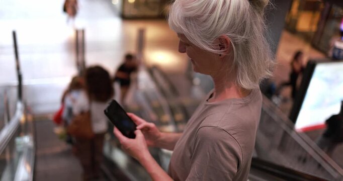 Caucasian senior woman using smartphone on escalator at train station - City lifestyle and technology concept - UGC content