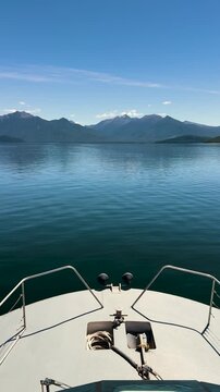 Vertical POV from boat's bow gliding across calm, glassy lake Manapouri toward distant mountains.