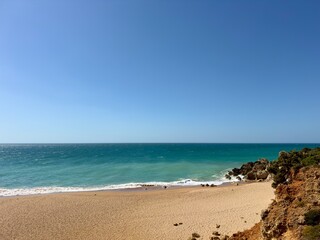 Obraz premium View of an empty bay with a sandy beach bordered by a rocky cliff and turquoise ocean water under a blue sky, paradise, tropical, holiday