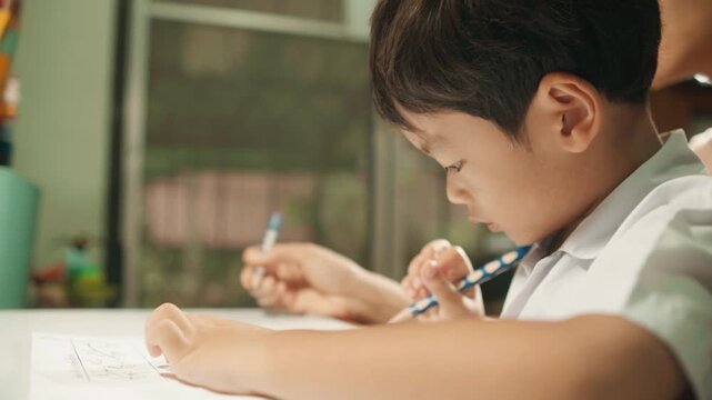 Footage of a young child practicing handwriting and completing an early-learning worksheet at a desk while a mother provides guidance and support beside him.