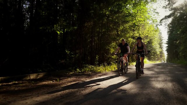 Cyclists riding together along a tranquil forest road at sunset, casting long shadows on the quiet rural path surrounded by tall green trees and soft golden light