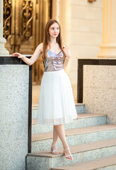 Fototapeta premium A young smiling woman poses elegantly against a backdrop of ancient historic buildings with golden Venetian style facades. The woman has long light brown hair and is wearing summer clothing.