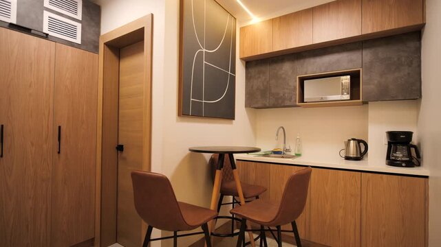Full framing of an interior space within an office: combined wooden furniture, preparation area with sink, microwave, coffee maker and kettle, high table with chairs and decorative abstract artwork.