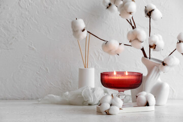 Red candle with cotton branches and aroma diffuser on a white textured background, cozy home decor concept, the arrangement creates a relaxing and warm ambiance