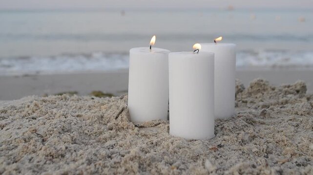 Three large paraffin white candles in sand on sandy beach of sea coast on background of sea waves. Flame fire. Candles burning on sand sea. Sea waves beach sand summer. Romantic mood romance. Sound