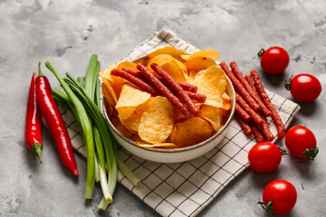 Fototapeta premium Bowl with delicious potato chips, jerky sticks, green onion, chili peppers and tomatoes on grey grunge background