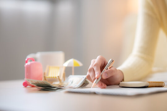 Close up woman writing travel budget beside cash, suitcase, beach chair, planning vacation expenses, saving money, personal finance, holiday fund, tourism concept.