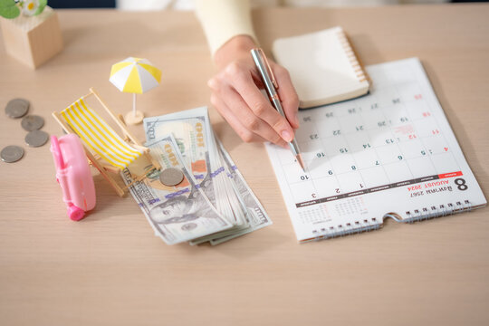 Top view woman marking calendar beside cash, coins, suitcase, beach chair, planning travel date, vacation budget, holiday savings, financial goal, tourism concept.