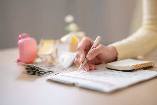 Close up hand marking calendar beside cash, notebook, suitcase, beach chair, planning vacation date, travel budget, holiday savings, financial planning concept.